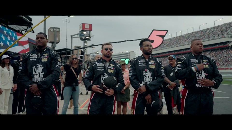 The Fallen Outdoors pit crew in matching black-and-red jumpsuits standing at attention on a packed NASCAR track, an American flag behind them.
