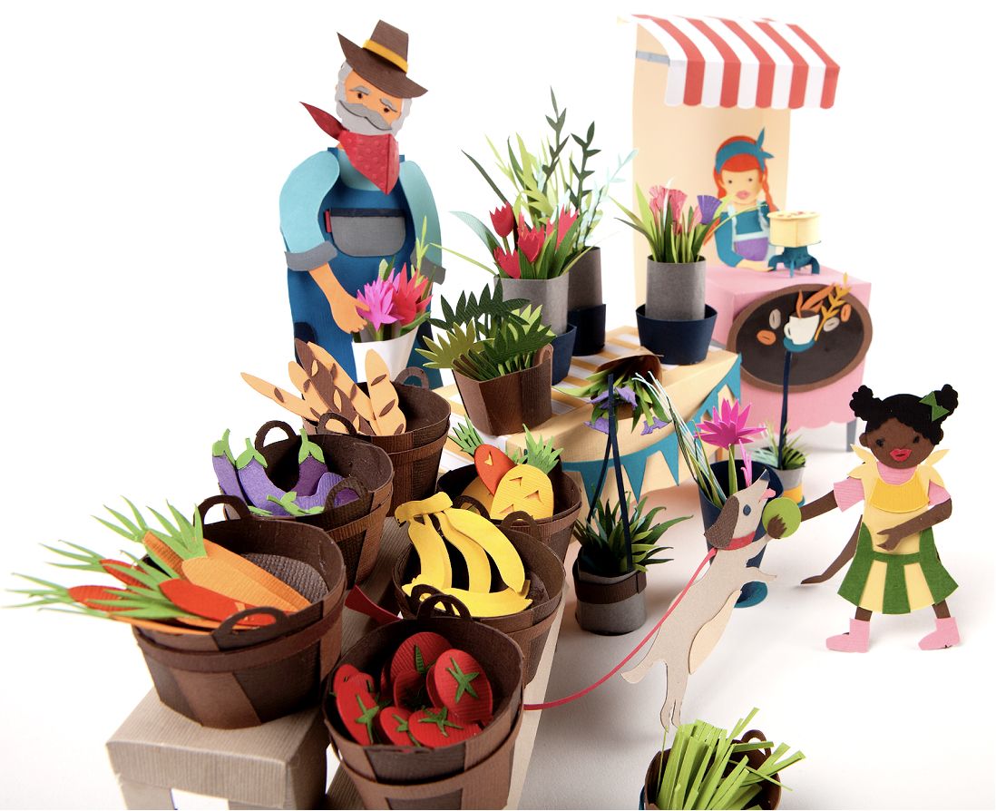 Three-dimensional paper craft sculpture of a farmers market scene: an elderly vendor in overalls and a cowboy hat arranges flowers beside barrels overflowing with paper-cut vegetables and fruit, while a young girl walks a dog in the foreground and a vendor tends a striped-awning coffee stand in the background.
